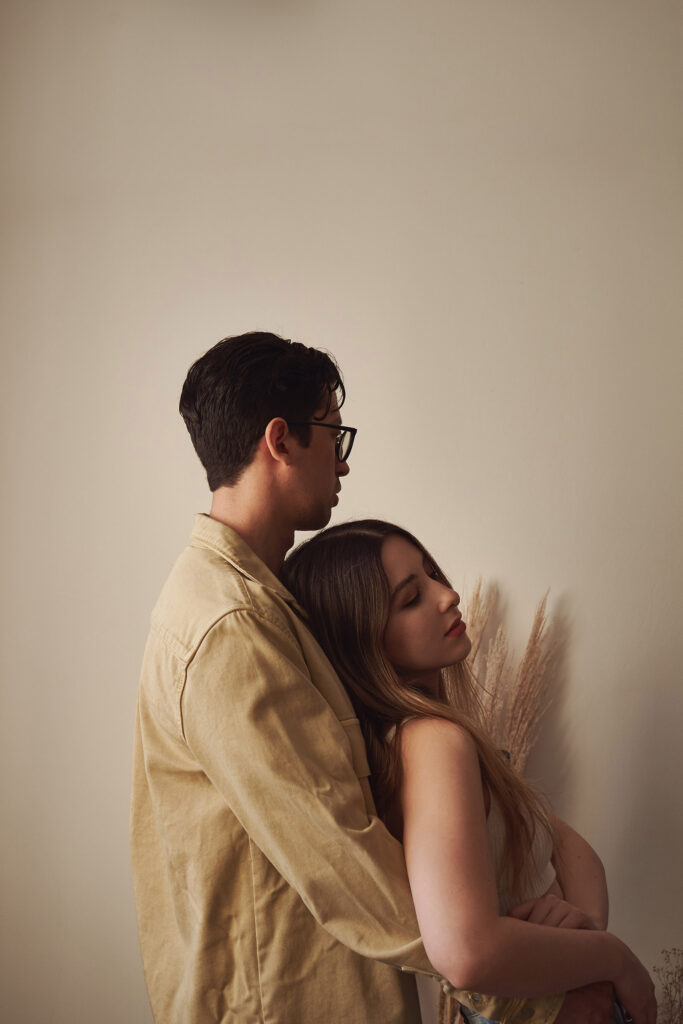 Gaby and Me a couple stand close together in a room with a beige wall. Gaby leans her head on My shoulder and he looks away from the camera. The ambiance is calm and intimate.